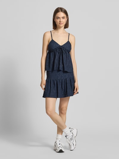 Modelka nosi Babydoll-Top mit Schößchendetail und Lochstickerei von REVIEW, mieszanka materiałów z dodatkiem streczu, krój regular fit, baskinka, haft angielski, długość tyłu w rozmiarze S: 27 cm, granatowy kolor, widoczne tekstury haftu, gładka powierzchnia, widok całej sylwetki z przodu.