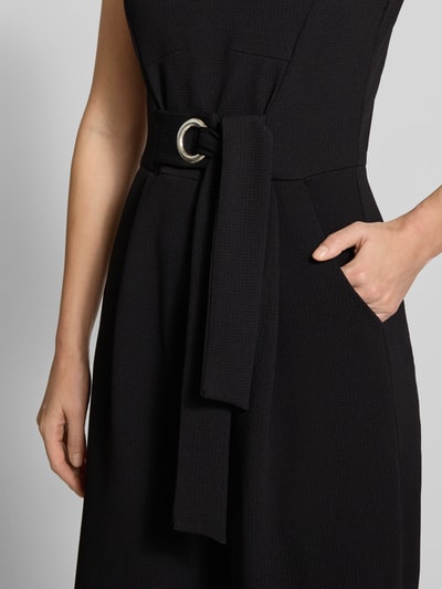 Kobiece model nosi Jumpsuit mit Rundhalsausschnitt od Apricot, czarny materiał z fakturą, pasek, kieszenie, zbliżenie na górną część stroju, detal.