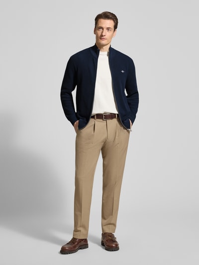 Männermodel trägt Cardigan mit Zweiwege-Reißverschluss und Label-Stitching von Gant aus reiner Baumwolle, Slim-Fit, Stehkragen, dunkelblau, kombiniert mit heller Hose, Ganzkörperansicht von vorne.