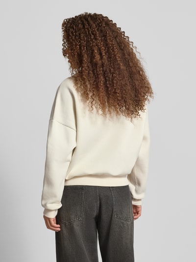 Modelka kobieca nosi Basic Sweater mit Rundhalsausschnitt und Bündchen am Ärmel od Review, kremowy, oversize, żebrowane wykończenia, haft z logo, długi rękaw, z opuszczonymi ramionami, widok całej sylwetki z tyłu.