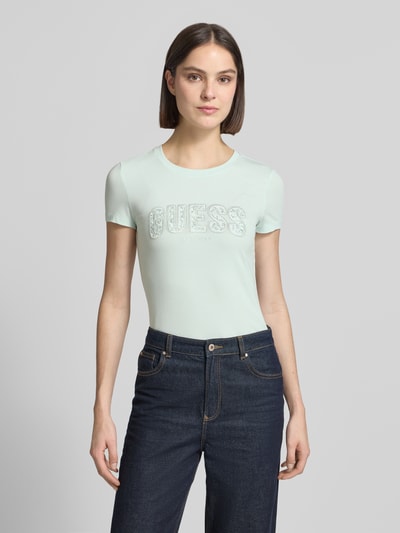 Frauenmodel trägt Slim Fit T-Shirt mit Strasssteinbesatz von Guess aus elastischem Baumwoll-Mix in Hellgrün, Rundhalsausschnitt, Logo-Detail, Rückenlänge bei Größe S 59 cm, Ganzkörperansicht von vorne.