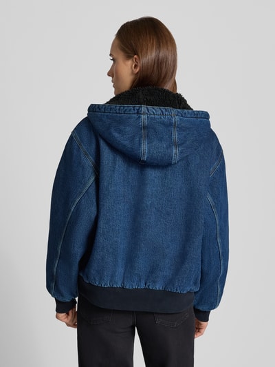 Frauenmodel trägt Jeansjacke mit Kapuze und Reißverschluss von Carhartt Work In Progress aus reiner Baumwolle in Dunkelblau, Ganzkörperansicht Rückansicht.