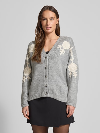 Frauenmodel trägt Strickjacke mit Knopfleiste von More & More, grau, V-Ausschnitt, gerippte Abschlüsse, Blumenmuster, Ganzkörperansicht von vorne.