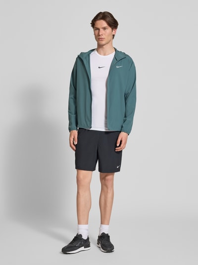Männermodel trägt Trainingsjacke mit Logo-Print und Stehkragen von NIKE TRAINING in Dunkelgrün mit Reißverschluss, seitliche Eingrifftaschen, elastische Ärmelabschlüsse, gerader Schnitt, Ganzkörperansicht von vorne.