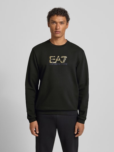 Männermodel trägt schwarzes Sweatshirt mit goldfarbenem Logo und Rundhalsausschnitt aus reiner Baumwolle, Regular-Fit, EA7 Emporio Armani, Ganzkörperansicht von vorne.