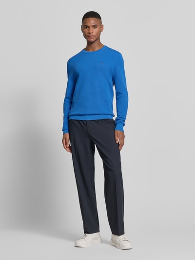 Mannenmodel draagt Regular Fit Strickpullover aus reiner Baumwolle van Tommy Hilfiger in effen blauw, gebreide look, logodetail, ronde hals, lange mouw, rompzoom geribd, puur katoen, ganzkörperansicht van voren.