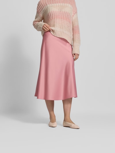 Frauenmodel trägt Midirock aus Satin mit Reißverschluss von Juvia in zartem Rosa, schmale Passform, Midi-Länge, Ganzkörperansicht von vorne.