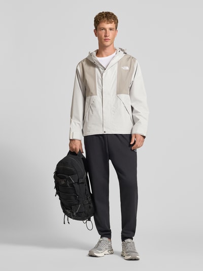 Męski model nosi kurtkę z kapturem Jacke mit Label-Stitching Modell 'REIGN ON 2.0' od The North Face, krój regular fit, jasnoszare odcienie, zamek błyskawiczny ukryty, Długość tyłu w rozmiarze M: 69 cm, Szerokość ramion w rozmiarze M: 52 cm, Szerokość klatki piersiowej w rozmiarze M: 63 cm, Długość rękawów: 65 cm, widok całej sylwetki od przodu.