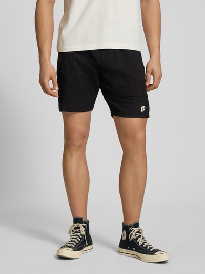 Männermodel trägt Shorts mit Label-Patch Modell 'LIBCO' von Pegador, Schwarz, Strukturmuster, Material-Mix, Gerader-Schnitt, Elastischer-Bund, Ganzkörperansicht von vorne.