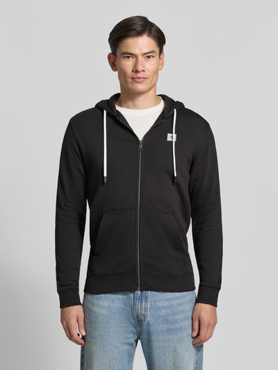 Männermodel trägt schwarze Sweatjacke mit Logo-Patch und Känguru-Tasche von Scotch & Soda, reine Baumwolle, Vorderansicht, Nahaufnahme Oberkörper.