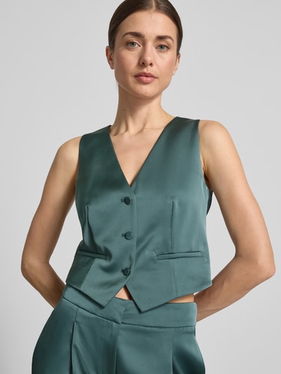 Frauenmodel trägt Weste aus Satin mit V-Ausschnitt von Jake*s Cocktail, smaragdgrün, mit Knopfleiste und Paspeltaschen, Nahaufnahme Oberkörper. Rückenlänge bei Größe 36: 40 cm, Slim-Fit.