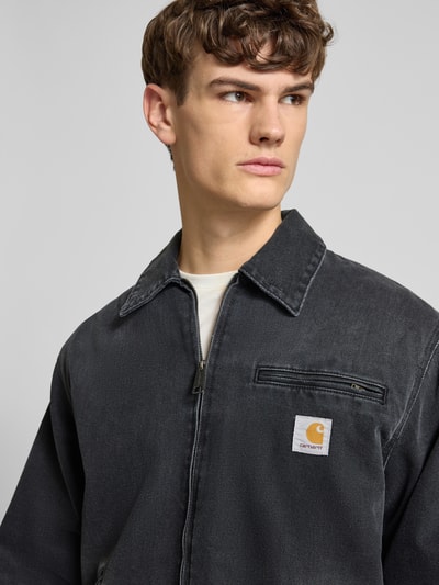 Männermodel trägt Jeansjacke mit Reißverschluss und Label-Detail Modell 'DETROIT' von Carhartt Work In Progress aus Baumwolle in Dunkelgrau, Nahaufnahme Oberkörper. Relaxed Fit, Umlegekragen, Eingrifftasche sichtbar.