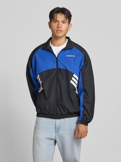 Männermodel trägt Regular Fit Trainingsjacke im Two-Tone-Design Modell 'MUC TT' von adidas Originals in Blau und Schwarz, Ganzkörperansicht von vorne. Herren Trainingsjacke von adidas Originals.