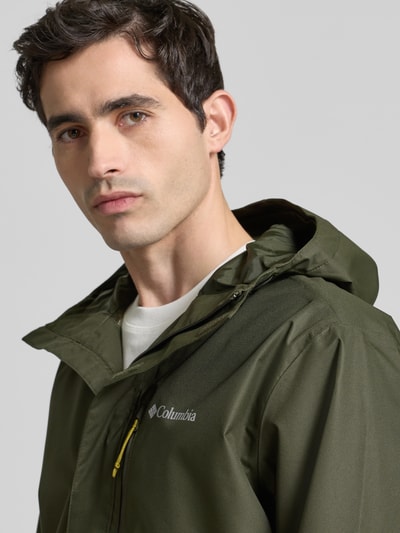 Männermodel trägt Jacke mit Kapuze Modell 'Hikebound™ II' von Columbia in Olivgrün, Regular-Fit, Logo, Nahaufnahme Oberkörper. Rückenlänge 74 cm, Schulterbreite 49 cm, Brustweite 61 cm, Ärmellänge 70 cm, Tunnelzug an Kapuze, Ärmelriegel, verdeckter Reißverschluss.