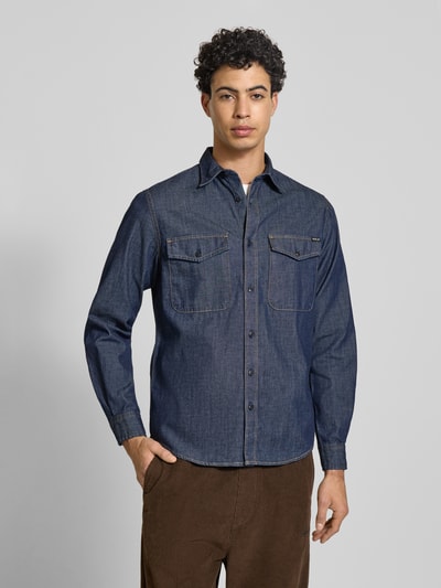 Męski model nosi Regular Fit Freizeithemd aus Baumwolle von Replay, ciemnoniebieska bawełna, detal z logo, krój regular fit, kołnierzyk typu kent, listwa guzikowa na całej długości, kieszenie na piersi z guzikiem, długość tyłu w rozmiarze M 75 cm, szerokość ramion w rozmiarze M 39 cm, długość rękawów 65 cm, widok całej sylwetki od przodu.