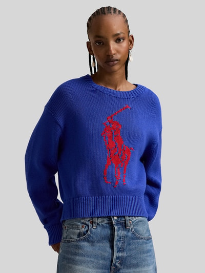 Frauenmodel trägt Regular Fit Strickpullover aus reiner Baumwolle von Polo Ralph Lauren in Two Tone Optik, blau mit rotem Intarsia-Logo, Crew Neck, langarm, gerippte Abschlüsse, Stricktextur, Nahaufnahme Oberkörper.