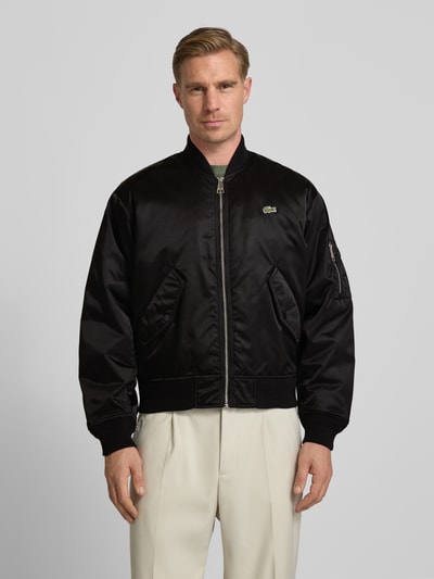 Männermodel trägt Regular Fit Bomberjacke aus Satin von Lacoste in Schwarz, gerippte Abschlüsse, Logo-Patch, Ganzkörperansicht von vorne.