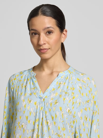 Frauenmodel trägt Regular Fit Bluse aus reiner Viskose mit Allover-Motiv-Print von Emily Van den Bergh in Hellblau mit gelben und weißen Herzmotiven und Tunika-Ausschnitt, weich fließende Textur, Langarm, elastische Ärmelabschlüsse; Damen Bluse, Nahaufnahme Oberkörper. Rückenlänge Größe 36: 68 cm, Ärmellänge Größe 36: 65 cm.