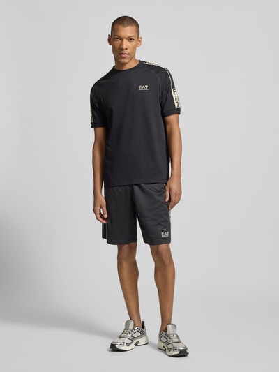 Męski model nosi T-shirt aus Viskose-Mix mit Logo von EA7 Emporio Armani, czarny, krój regular fit, okrągły dekolt, długość tyłu 70 cm, Ganzkörperansicht von vorne.
