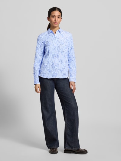 Modelka nosi HBK Zümdholzstreifen Streifen Bluse mit Blumen Applikationen von Christian Berg Woman, jasnoniebieskie paski, kwiatowe aplikacje, luźny krój, kołnierzyk koszulowy, bawełna, długość tyłu 62 cm, długość rękawa 60 cm, widok całościowy z przodu.
