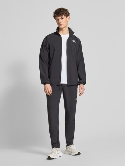 Męski model nosi Regular Fit Jacke mit Stehkragen Modell 'Mountain' od The North Face, czarna elastyczna mieszanka materiałów, nadruk z logo, zamek błyskawiczny, wpuszczane kieszenie, elastyczne wykończenia. Ganzkörperansicht von vorne.