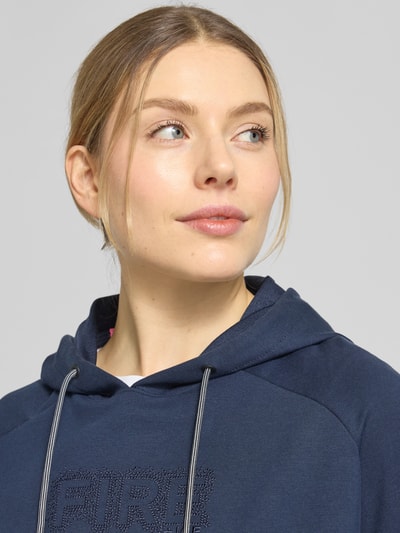 Modelka kobieta ma na sobie T-Shirt aus Baumwoll-Mix mit Kapuze Modell 'SHARON3' von FIRE + ICE w kolorze granatowym, z logo, bawełniano-elastanowy materiał, krótki rękaw, kaptur z troczkiem, luźny krój, detal marki, zbliżenie na górną część ciała.