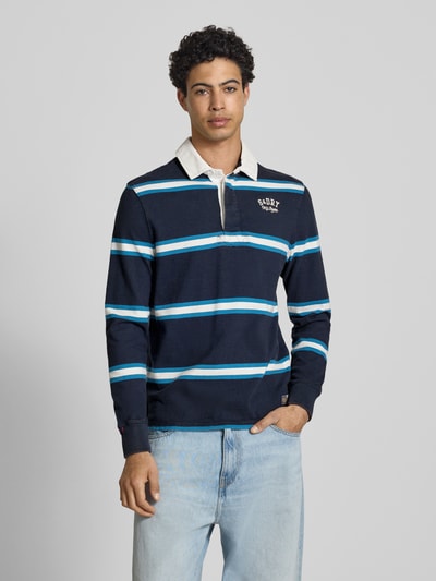 Männermodel trägt Regular Fit Poloshirt mit Logo-Stitching von Superdry in Dunkelblau mit weißen und blauen Streifen, reine Baumwolle, Polokragen, kurze Knopfleiste, Label-Details, Ganzkörperansicht von vorne.