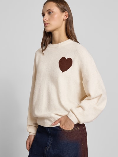 Vrouwenmodel draagt Regular Fit pullover in tricotlook van REVIEW, materiaalmix met stretch, beige met bruin hartmotief, lange mouw, ribboorden, zichtbare bies, overlappende schouders, voorkant volledig zichtbaar, Ganzkörperansicht von vorne.