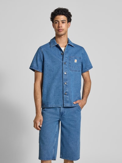 Męski model nosi Regular Fit Jeanshemd mit Brusttasche Modell 'SUN' od Blend, czysta bawełna, niebieski kolor, kołnierzyk typu kent, listwa guzikowa, naszywka z logo, brustasza, długość tyłu w rozmiarze M: 70 cm, szerokość ramion w rozmiarze M: 42 cm, długość rękawów w rozmiarze M: 23 cm, tekstura jeansowa, widok całej sylwetki z przodu.