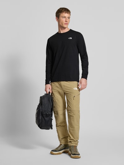 Męski model nosi Bluzka z długim rękawem z wyhaftowanym logo od The North Face z mieszanki bawełny, krój regular fit, okrągły dekolt, długość tyłu w rozmiarze M: 70 cm, kolor czarny, gładka faktura, widok całej sylwetki z przodu.