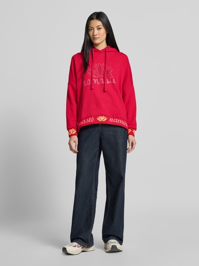 Kobiece modelka nosi Oversized Hoodie aus reiner Baumwolle Modell 'Namasté' von miss goodlife, kolor czerwony, nadruk z napisem, obszycie z kamieni stras, elastyczne wykończenia, szeroki krój, całkowity widok z przodu
