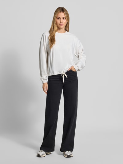 Frauenmodel trägt Relaxed Fit Langarmshirt aus reine Baumwolle von DRYKORN, weiß, gerippte Blende und Bündchen, Tunnelzug im Saum, schwarze Hose, Ganzkörperansicht von vorne.