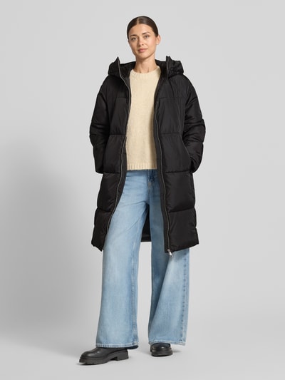Frauenmodel trägt Daunenmantel mit Kapuze Modell 'C_Polliana' von BOSS Orange, unifarben schwarz, Longline, mit Zweiwege-Reißverschluss, seitlichen Eingrifftaschen und Raglanärmeln, gerippte Ärmelabschlüsse, strukturiert durch Nähte, Ganzkörperansicht von vorne.