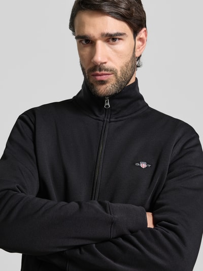 Männermodel trägt Regular Fit Sweatjacke mit Stehkragen Modell 'Gield' von Gant in Schwarz, Logo-Stitching, Nahaufnahme Oberkörper. Baumwoll-Mix, Reißverschluss, elastische Abschlüsse.