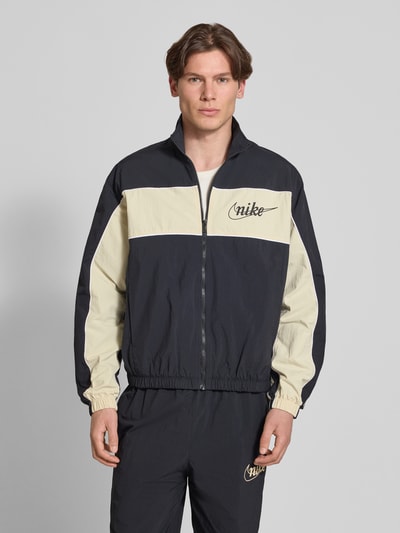Männermodel trägt Trainingsjacke mit Logo und Reißverschluss von Nike in Schwarz und Beige, Regular-Fit, Stehkragen, Ganzkörperansicht von vorne. Rückenlänge bei Größe M: 67 cm, Ärmellänge bei Größe M: 64 cm.