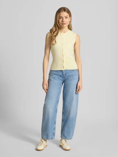 Vrouwenmodel draagt Strickweste aus Viskose-Mix mit Rundhalsausschnitt van Samsøe Samsøe in lichtgeel, regular fit, model 'ANNE', knopsluiting, V-hals, gladde textuur, gecombineerd met lichtblauwe jeans en sneakers, volledige lichaamsweergave van voren.