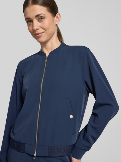 Kobiece modelka nosi kurtkę Jacke mit Zweiwege-Reißverschluss Modell 'Junipa' od JOOP!, mieszanka materiałów z dodatkiem streczu, krój regular fit, stójka, kolor granatowy, szerokość w rozmiarze 36: 52 cm, długość tyłu: 52 cm, długość rękawów: 62 cm, zamek błyskawiczny dwustronny, widok na górną część ciała z przodu
