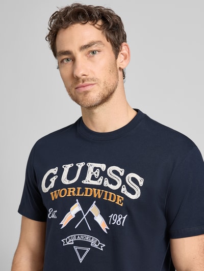 Męski model nosi T-Shirt mit Rundhalsausschnitt und Label-Stitching od Guess, czysta bawełna, krój regular fit, model 'FLAGS AND CORD', kolor granatowy, nadruk i naszywka z przodu, zbliżenie na górną część ciała