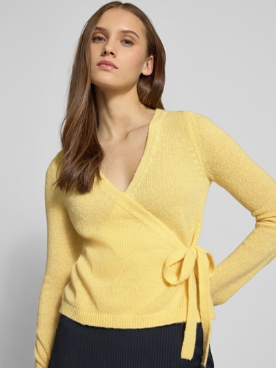 Vrouwenmodel draagt Strick-Cardigan in wikkellook van Review, regular fit, V-hals, lange mouwen, striksluiting, ruglengte 50 cm, mouwlengte 66 cm, gele kleur, fijngebreide structuur, Nahaufnahme bovenlichaam.