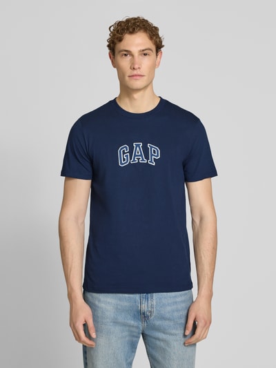 Männermodel trägt T-Shirt mit Logo-Patch und Rundhalsausschnitt von GAP in Dunkelblau, gerade Passform, reine Baumwolle, Ganzkörperansicht von vorne.
