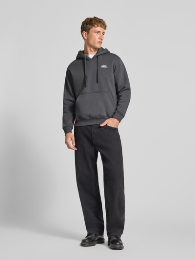 Mężczyznamodel nosi Hoodie mit Label-Details od Alpha Industries, mieszanka bawełny, luźny krój, kaptur z tunelem, kieszeń kangurka, prążkowane wykończenia, długość tyłu w rozmiarze M: 69 cm, długość rękawów w rozmiarze M: 70 cm, ciemnoszary, gładka faktura, widok całej sylwetki z przodu
