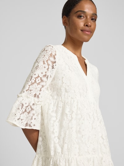 Frauenmodel trägt Vero Moda Loose Fit Spitzenkleid aus Baumwoll-Mix Modell 'HONEY' in Optik: Unifarben mit Spitzen-Muster, V-Ausschnitt, 3/4-Arm und Glockenärmel, Spitze-Textur, knielang, Nahaufnahme Oberkörper.