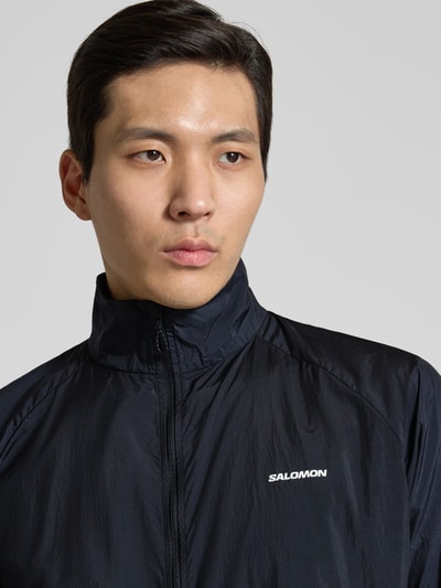 Männermodel trägt Jacke mit Stehkragen und Logo von Salomon in Schwarz, Regular-Fit, strukturierte Oberfläche, Nahaufnahme Oberkörper. Rückenlänge bei Größe M: 70 cm, Schulterbreite: 42 cm, Brustweite: 61 cm, Ärmellänge: 66 cm, Reißverschluss.