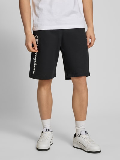 Männermodel trägt schwarze Shorts mit Logo-Print und -Stitching von CHAMPION, Baumwoll-Mix, lockerer Schnitt, Nahaufnahme Unterkörper.