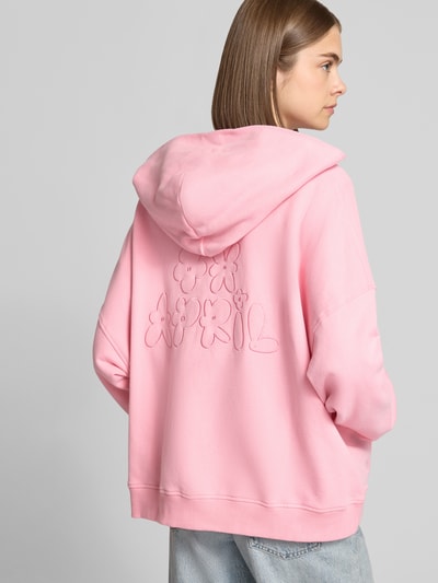 Młodzieżowy model nosi Oversized Sweatjacke mit Zweiwege-Reißverschluss Modell 'Flower' von OH APRIL z bawełny, różowy kolor, bluza z kapturem, kieszeń kangurka, krój oversized, haft kwiatowy z tyłu, widok na cały tył.