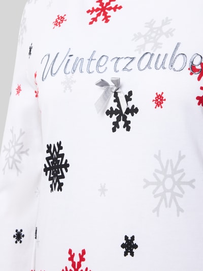 Frauenmodel trägt Nachthemd aus reiner Baumwolle Modell 'Winterzauber' von LOUIS & LOUISA, weiße Grundfarbe mit roten, schwarzen und grauen Schneeflocken, taillierte Passform, Motiv-Stickerei, Rundhalsausschnitt, langärmliges Design, Nahaufnahme Oberkörper.