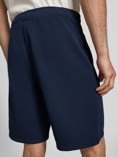 Männermodel trägt Bermudas mit elastischem Bund und Label-Stitching von CHAMPION aus Baumwoll-Mix in Dunkelblau, Längsnähte, Regular-Fit, Ganzkörperansicht Rückansicht.