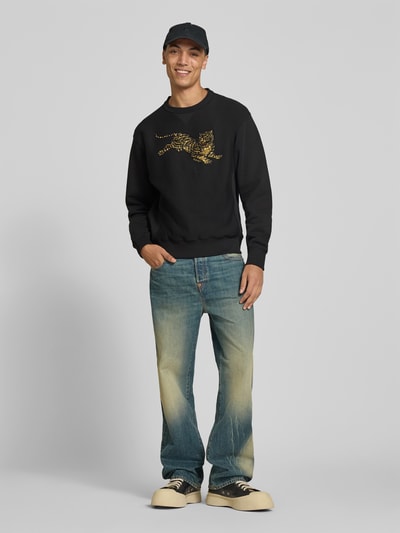 Mannenmodel draagt sweatshirt uit puur katoen van Kenzo, recht pasvorm, opstaande kraag, motiefprint met pailletten, ribboorden, ruglengte bij maat M 66 cm, zwart, met goudkleurig motief, blauwe jeans, sneakers, geheelvooraanzicht.