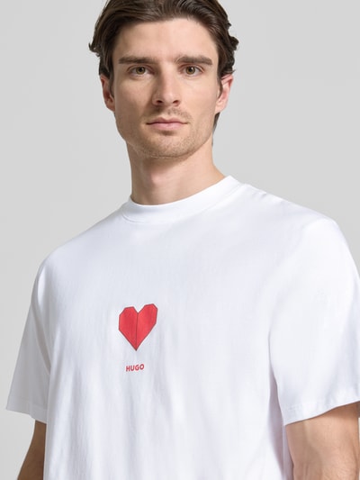 Männermodel trägt Regular Fit T-Shirt aus reiner Baumwolle Modell 'DALENTIN' von HUGO in Weiß mit rotem Herz-Print und Label, Nahaufnahme Oberkörper.