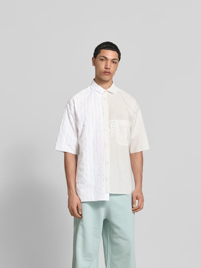 Männermodel trägt Regular Fit Freizeithemd aus Baumwolle von MM6 Maison Margiela im Asymmetrischer-Look, Halbarm, Kent- und Button-Down-Kragen, Unifarbenes-Design mit feiner Struktur, Rückenlänge 79 cm, Schulterbreite 48 cm, Ärmellänge 30 cm, Ganzkörperansicht von vorne.